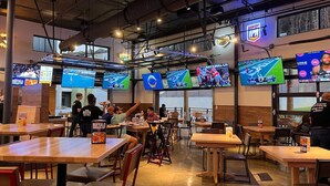 Un restaurant doté de plusieurs écrans diffusant des événements sportifs, de tables et de chaises en bois, ainsi que d’un comptoir où le personnel sert les plats.