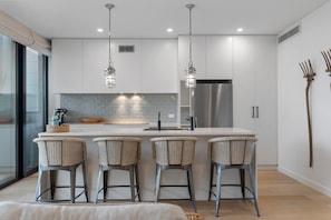 A modern kitchen with white cabinets, stainless steel appliances, wicker bar stools, and pendant lighting.
