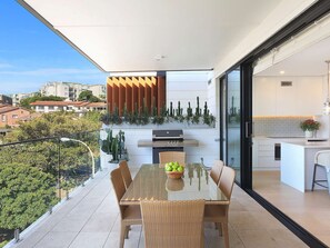 Un balcón moderno con una mesa de vidrio, sillas de mimbre, una parrilla y vistas a la ciudad.