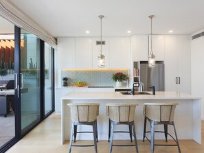 A modern kitchen with a central island, bar stools, pendant lighting, and a view through a glass door. - Pine Tree Penthouse (Terrigal)