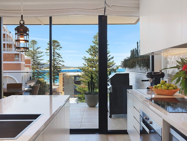 A modern kitchen with a view of a pool and buildings, a hanging light fixture, and a bowl of green apples.