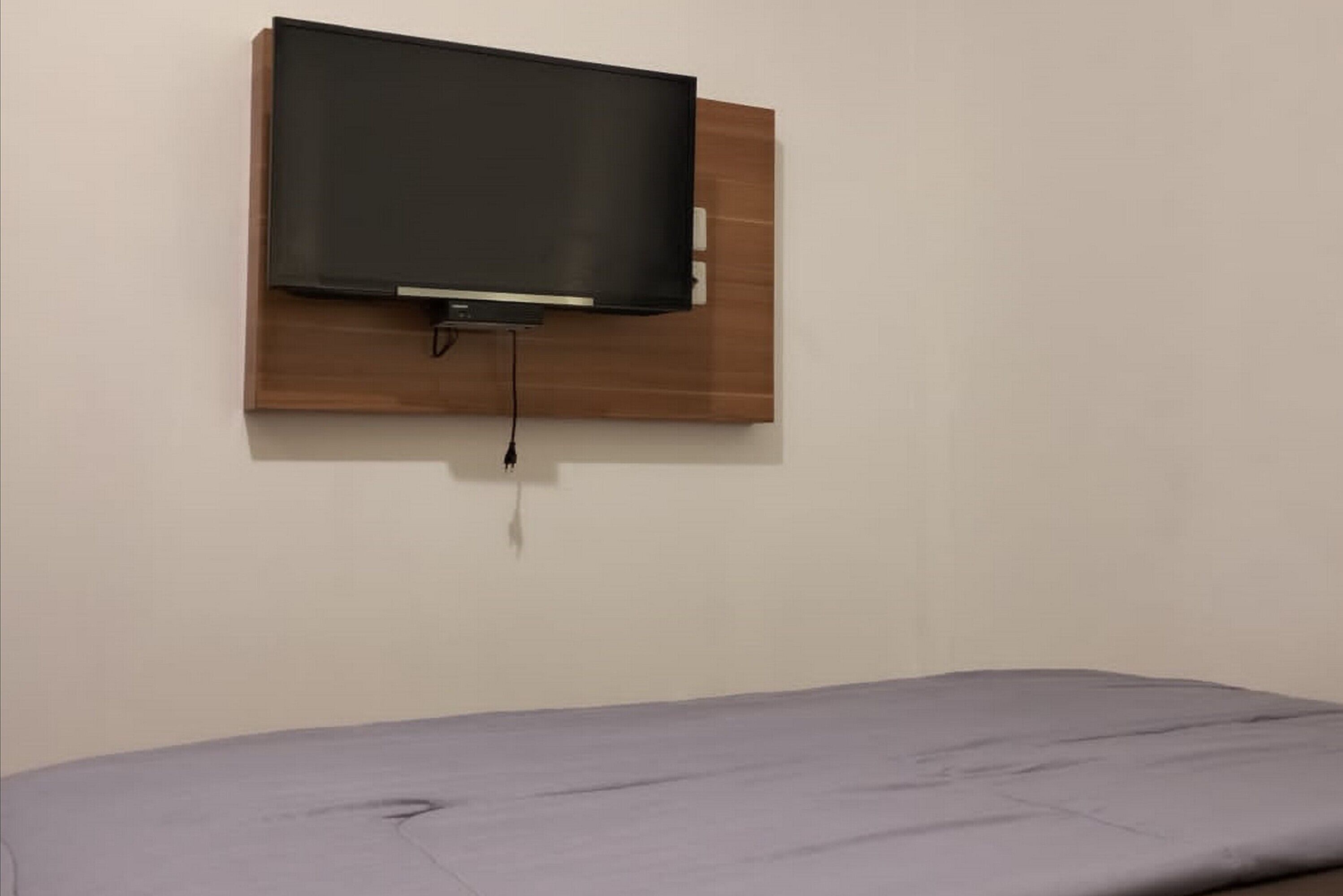 A room with a mounted television on a wooden panel and a bed with a grey mattress.