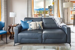 A modern living room with a dark leather sofa, a glass coffee table, and a view of mountains through large windows.