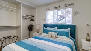 A bedroom with a bed, a nightstand, a bedside table, a window with blinds, a closet with shelves, and a wall decoration.