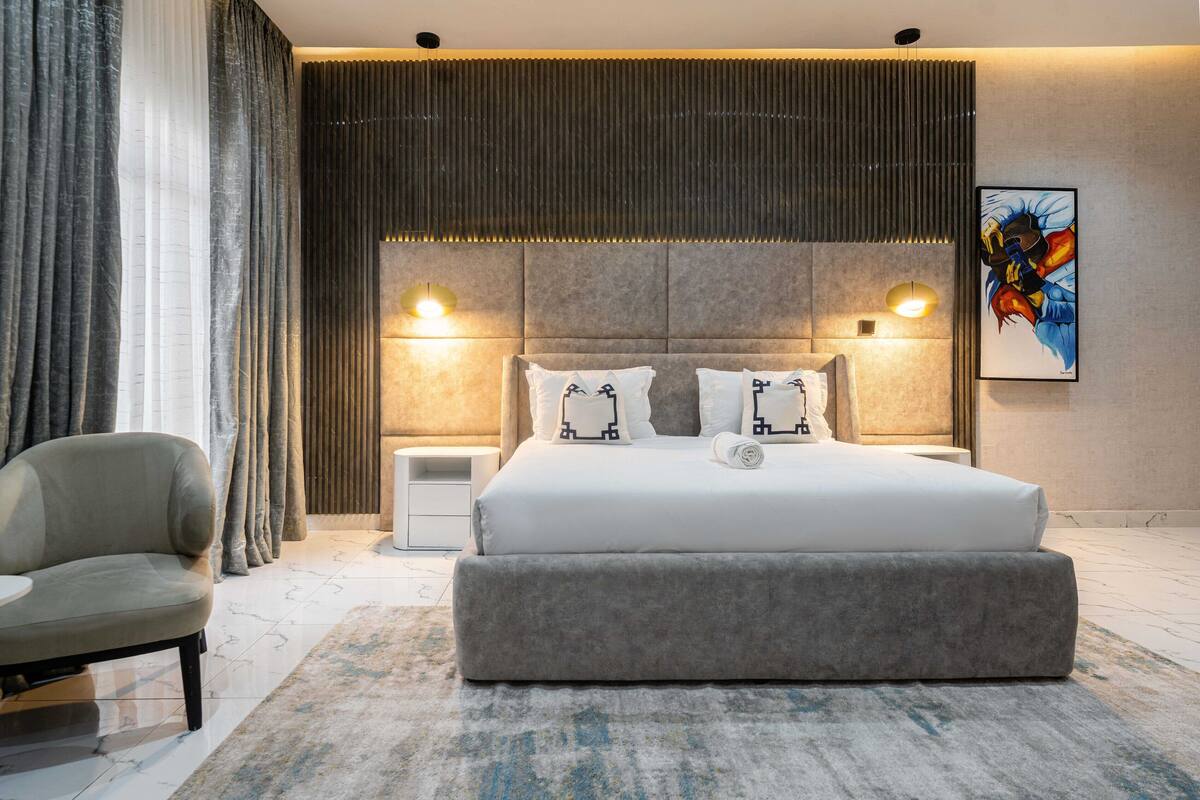 A modern bedroom with a large bed, two bedside lamps, a grey upholstered chair, and a nightstand.