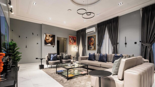 A modern living room with a beige sofa, a glass coffee table, and a black side table. There are two paintings on the wall, a potted plant, and a large mirror. - George Residence Parkview (Lagos)