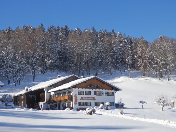 Un chalet enneigé, avec une façade en bois et un balcon, entouré d’arbres chargés de neige.