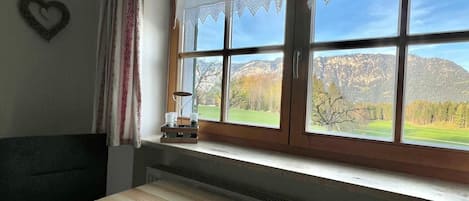 A room with a wooden table, a window with lace curtains, and a view of mountains.