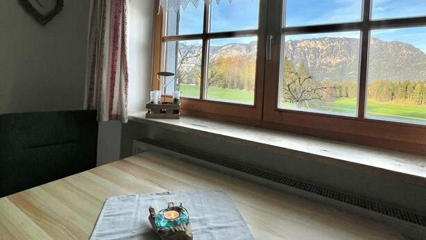 Une pièce avec une table en bois, une fenêtre ornée de rideaux en dentelle, un décor en forme de cœur sur le mur et une vue sur les montagnes.