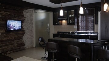 Una cocina moderna con un muro de acento de piedra, un televisor sobre una zona de bar, y un amplio ventanal con persianas.