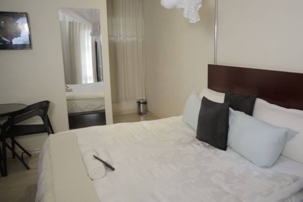 A hotel room with a large bed, a small table, a chair, a mirror, and a window with curtains.