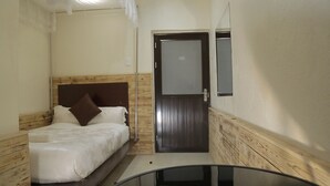 A small, narrow hotel room with a single bed, a wooden headboard, a door with a frosted glass panel, and a small table in the corner. - Commonlens Hotel (Entebbe)