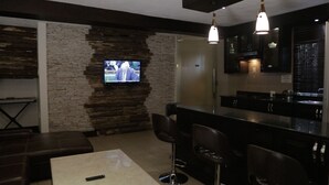 A modern living room with a stone accent wall, a flat-screen TV, a bar area with stools, and a kitchen visible through the glass partition. - Commonlens Hotel (Entebbe)