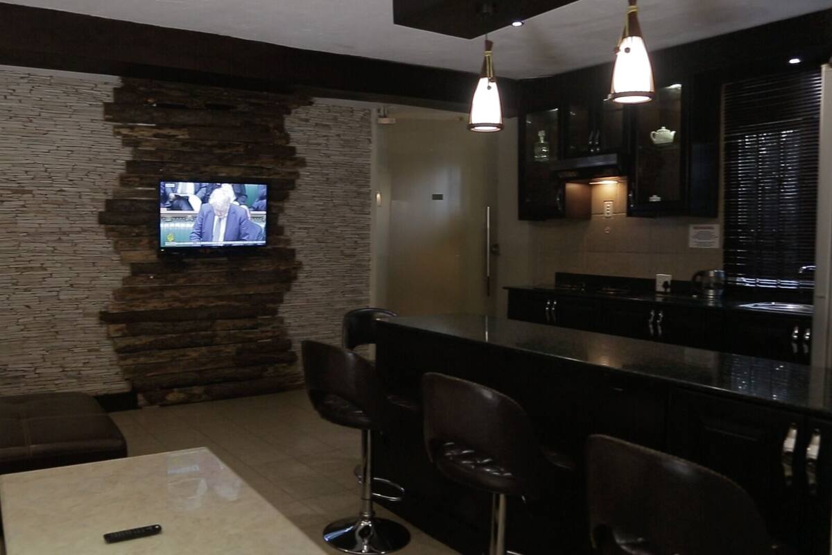 A modern living room with a stone accent wall, a flat-screen TV, a bar area with stools, and a kitchen visible through the glass partition.