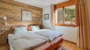 Un dormitorio con cabecera de madera, cama con varios almohadones, mesita de noche con lámpara, ventana con cortinas y vista a zonas verdes.