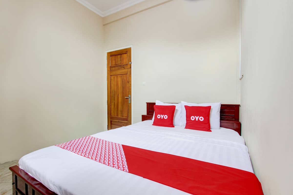 Una cama bien hecha con una colcha blanca y roja, dos almohadas con la palabra "OYO" y una puerta de madera al fondo.