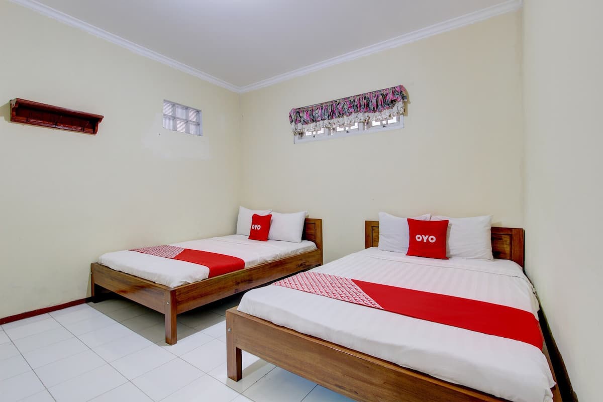 Una habitación de hotel con dos camas de madera, cada una con ropa de cama blanca y detalles rojos, una ventana pequeña con persianas, un estante de madera y una cortina decorativa.