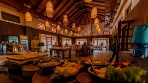 A buffet area with various food items and a person serving. There are hanging lights and wooden beams on the ceiling. - Banbas Resort, Chitwan (Sauraha)