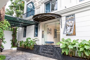 Un edificio moderno con un acceso cubierto, una escalera que lleva a la entrada y abundante vegetación alrededor.
