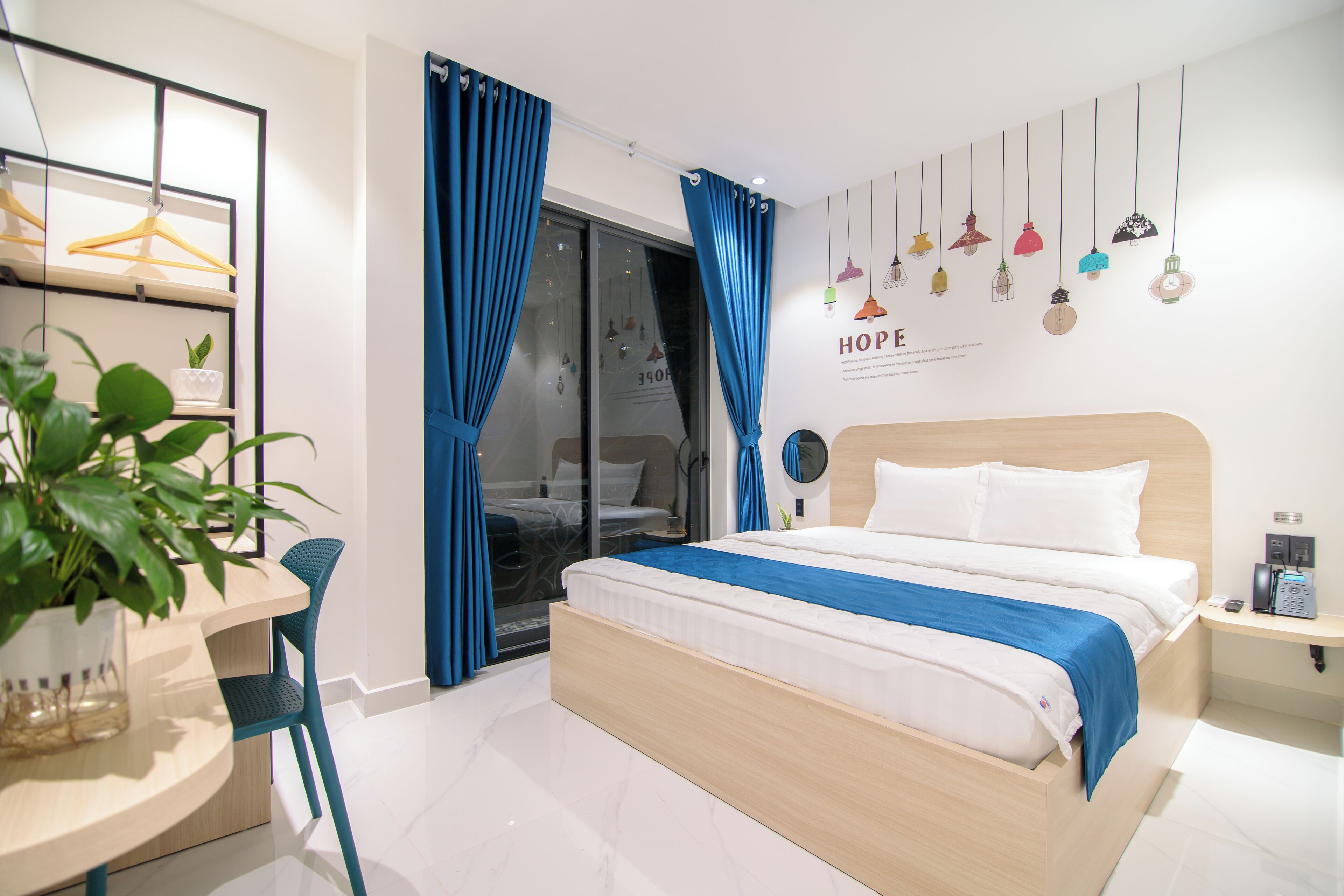 Un dormitorio moderno con una cama, un escritorio y una silla. Hay una planta en el escritorio, y la habitación tiene un ventanal con cortinas.