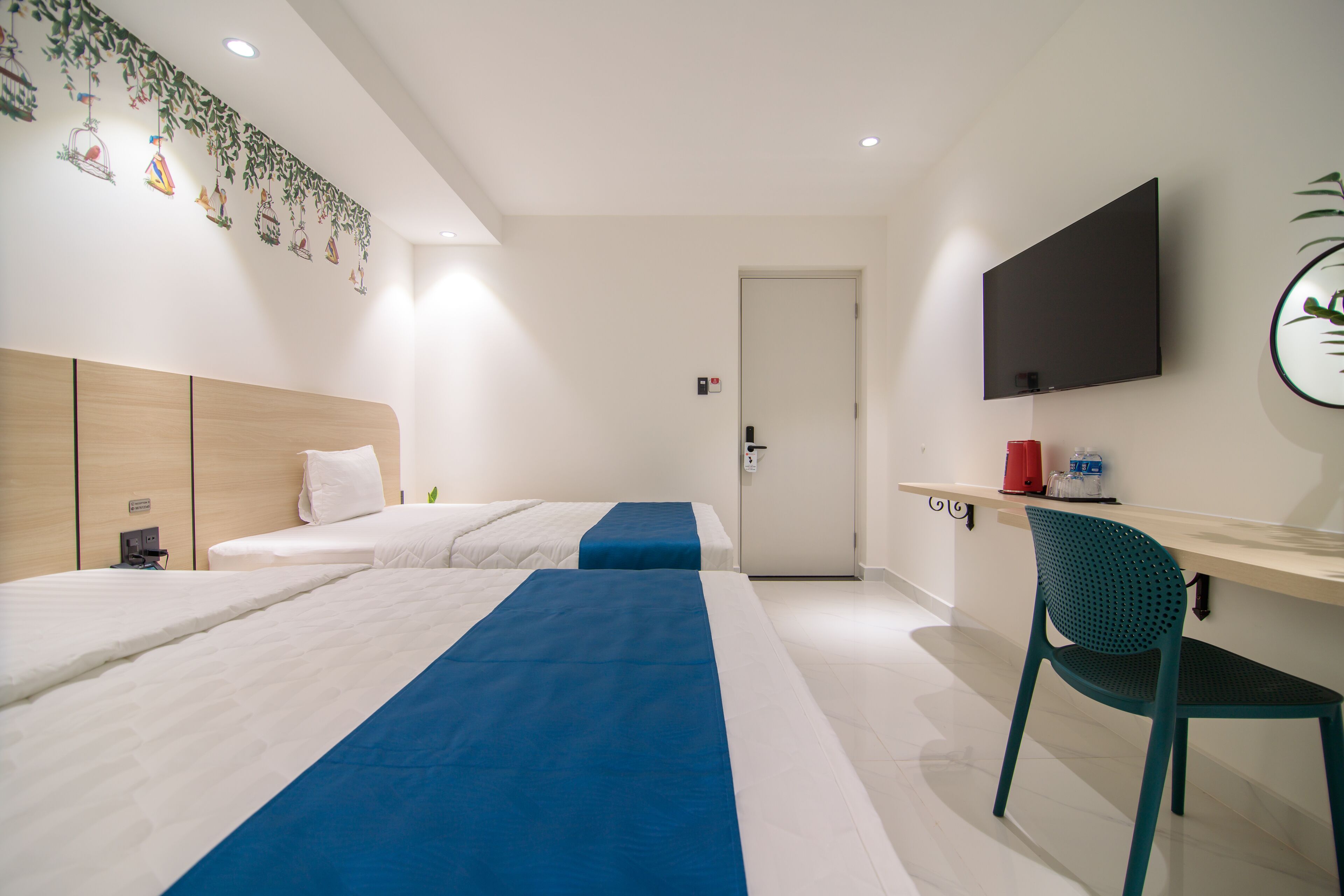 A hotel room with two beds, a desk with a chair, a flat-screen TV, and a door.