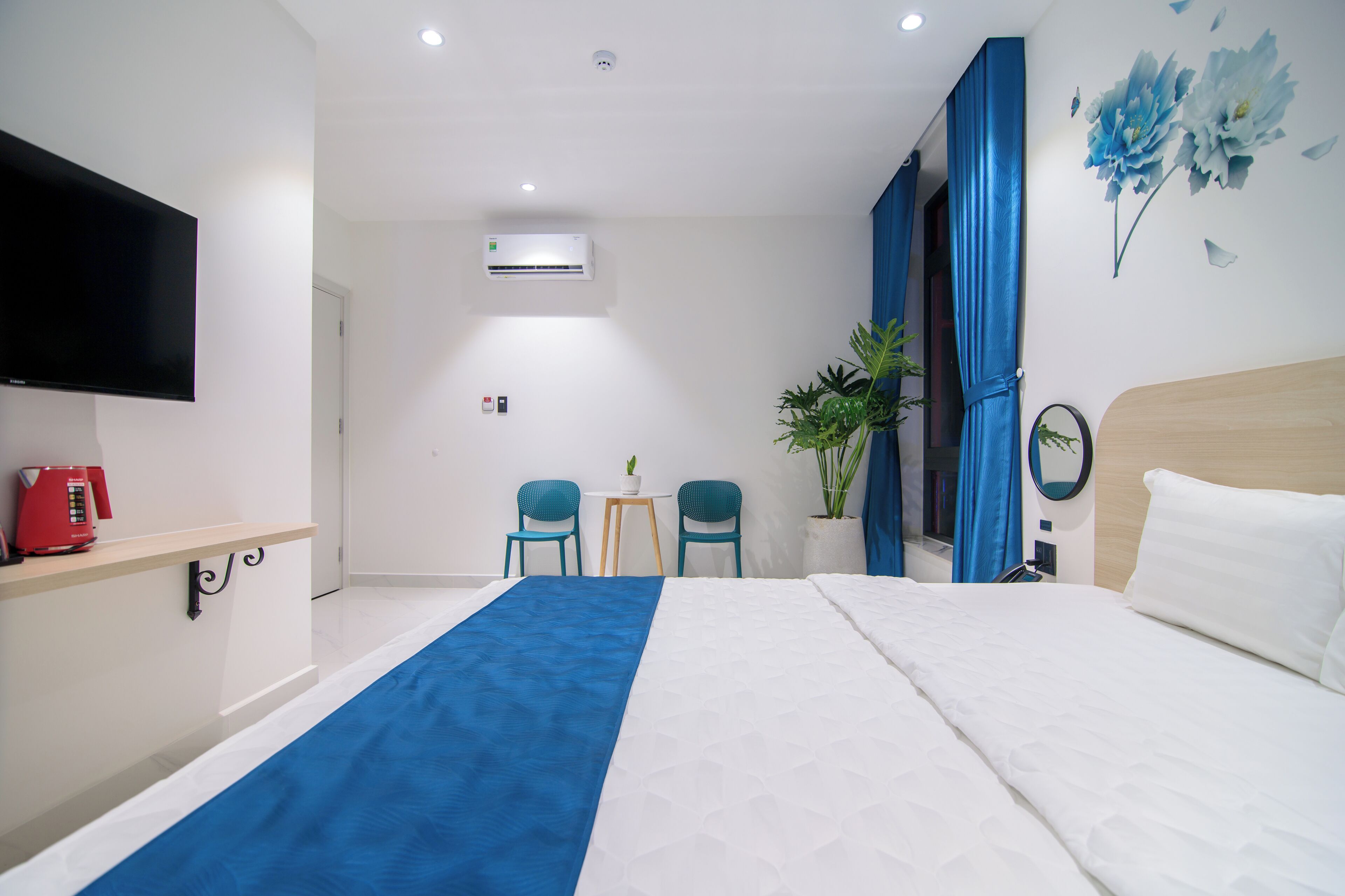 Ein Hotelzimmer mit einem Bett, einem Fernseher, einem Schreibtisch mit rotem Kaffeeautomaten, zwei Stühlen, einem kleinen Tisch mit einer Pflanze und einer Wandklimaanlage.