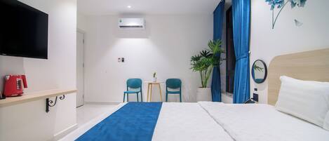 A hotel room with a bed, a TV, a desk with a red coffee maker, two chairs, a small table with a plant, and a wall-mounted air conditioner.