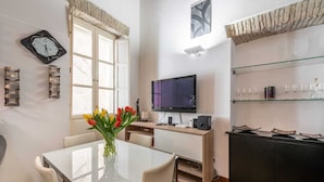 A modern living room with a glass dining table, a flat-screen TV, a wooden desk, and a black cabinet with shelves.