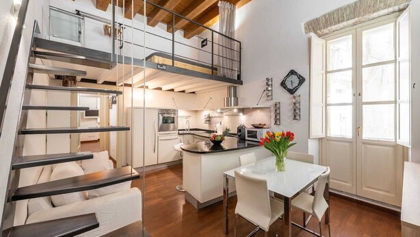 A modern kitchen with a central island, a dining area with a white table and chairs, and a staircase leading to a loft bedroom.