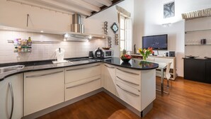 A modern kitchen with white cabinets, a black countertop, and a stainless steel range hood. There is a dining area with a table and chairs, and a flat-screen TV mounted on the wall.