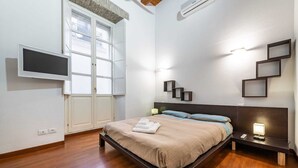A bedroom with a bed, bedside table, wall-mounted TV, and a window with blinds.
