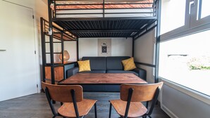 Quarto compacto com cama beliche, sofá, duas cadeiras, mesa de madeira e janela grande.