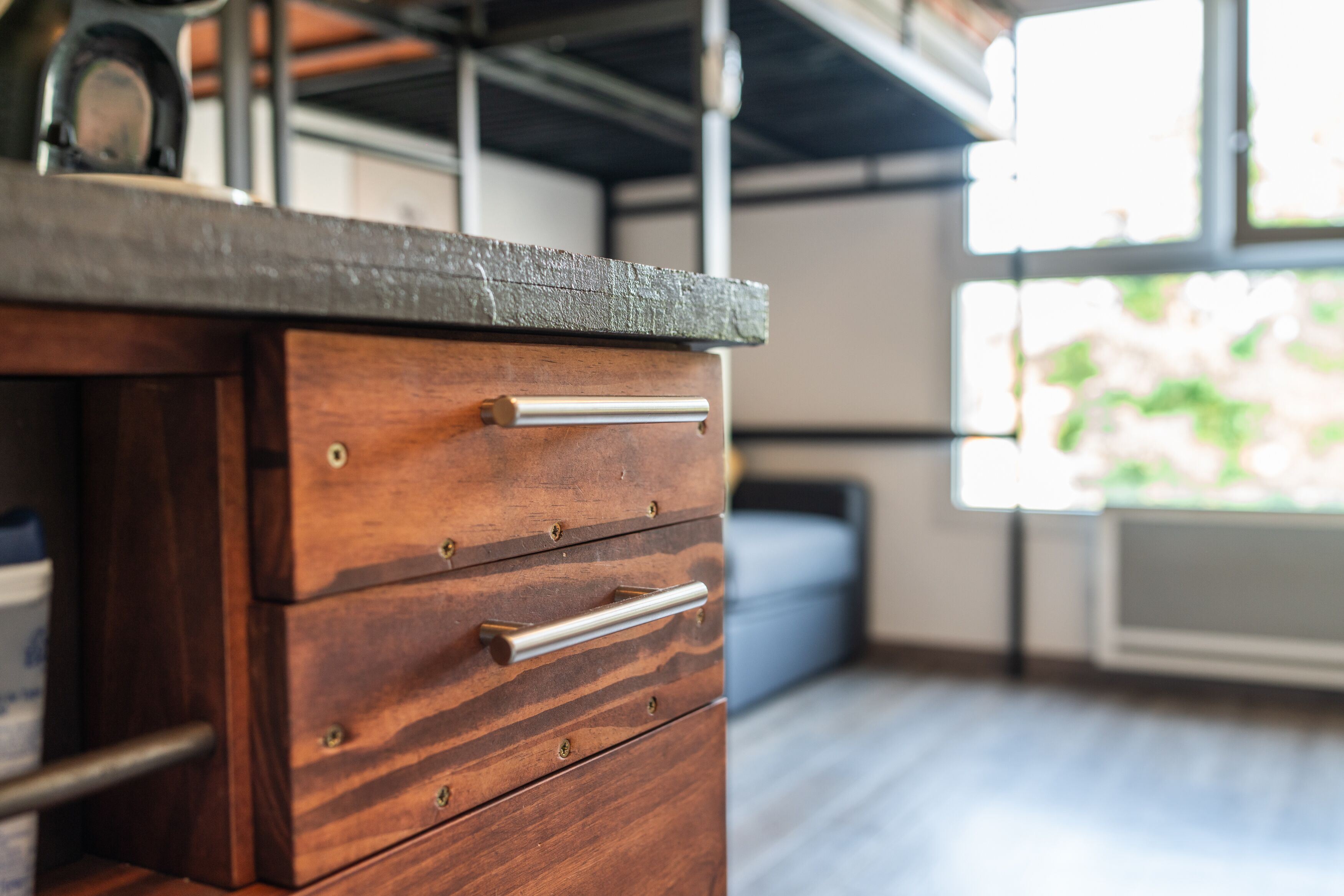 A wooden dresser with drawers and a metal handle, a window with a view of greenery, and a metal shelf with a clock.