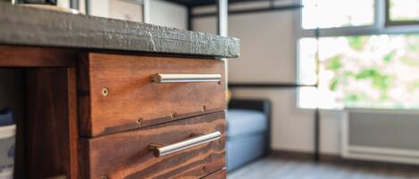 Un aparador de madera con cajones y un tirador metálico, una ventana con vista a follaje, y una estantería metálica con un reloj.