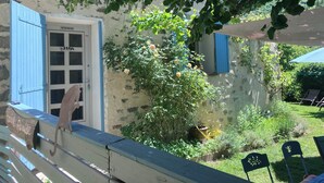 Un edificio rústico con una puerta azul, un banco de madera con un cartel, y un jardín con una planta en maceta y una silla.