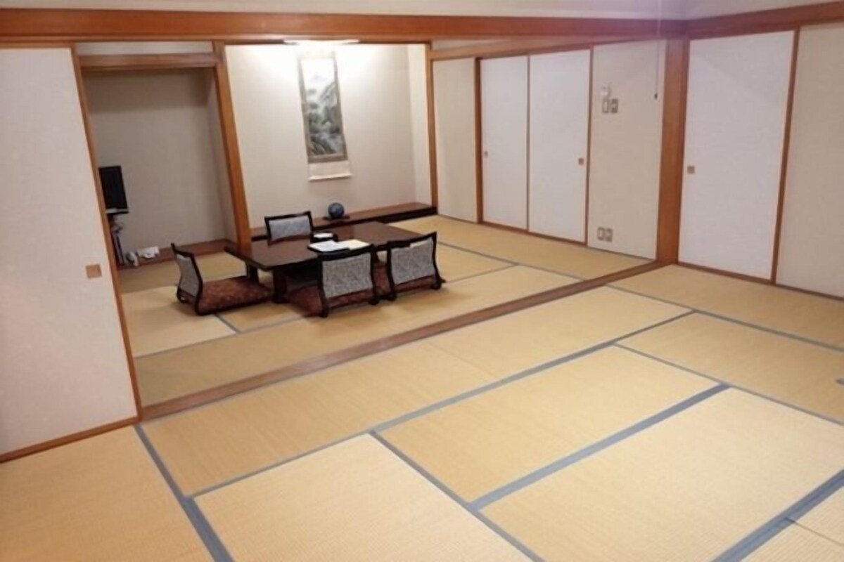 Um espaço com tatames, uma mesa baixa, cadeiras, portas deslizantes e um quadro na parede.