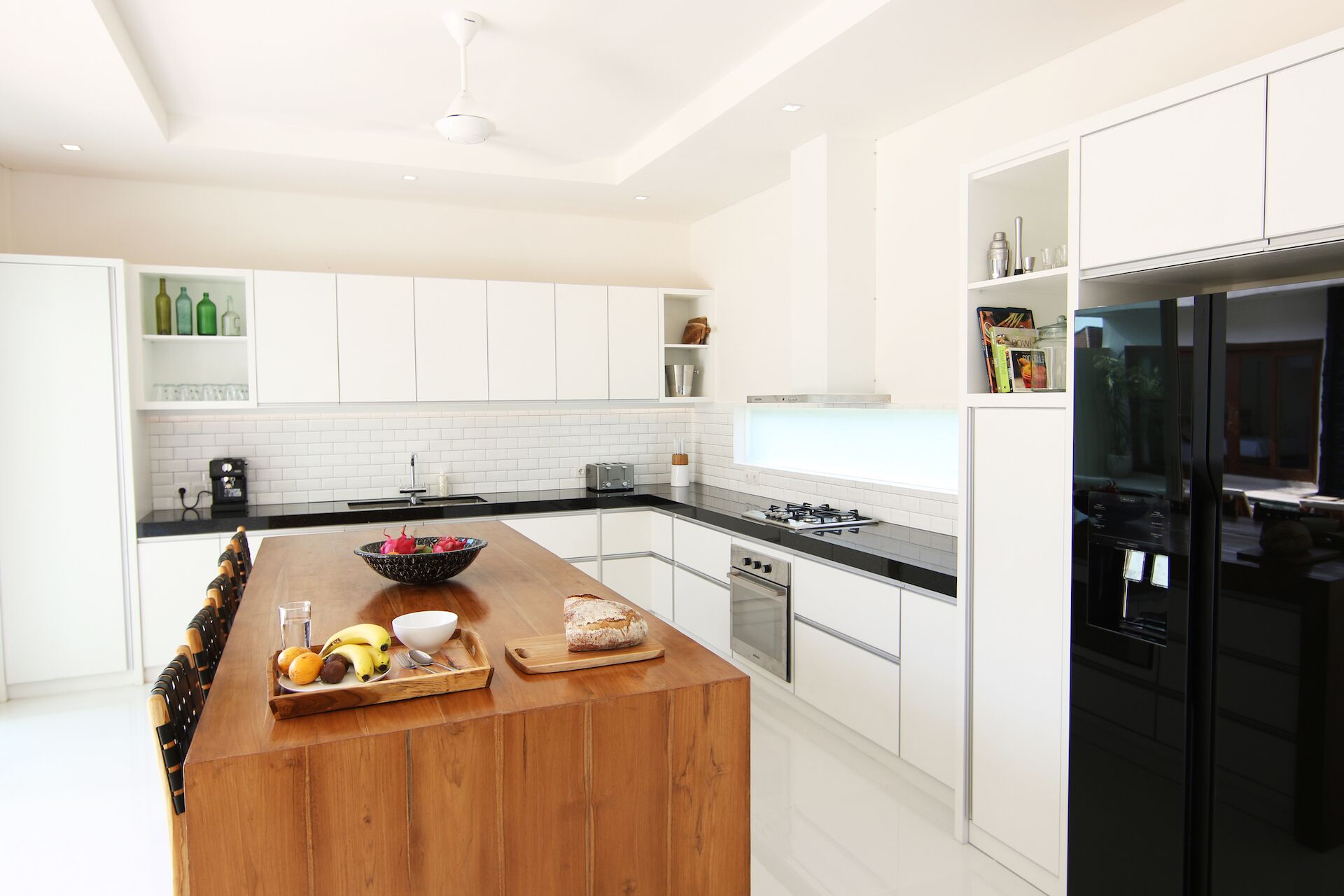 A modern kitchen with white cabinetry, a wooden island with fruits and bread, a black refrigerator, and a window overlooking the room.