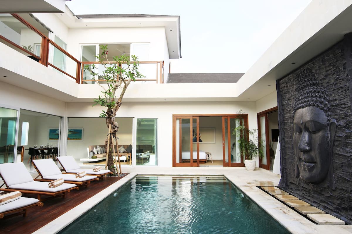 A modern indoor pool area with lounge chairs, a Buddha statue, and a dining area with a view of the pool.