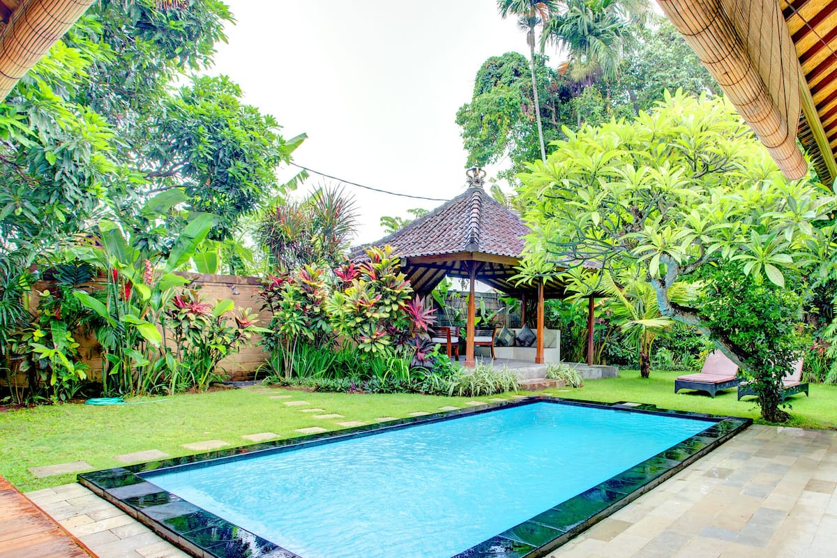 A swimming pool with a tiled deck, surrounded by lush greenery and a gazebo.