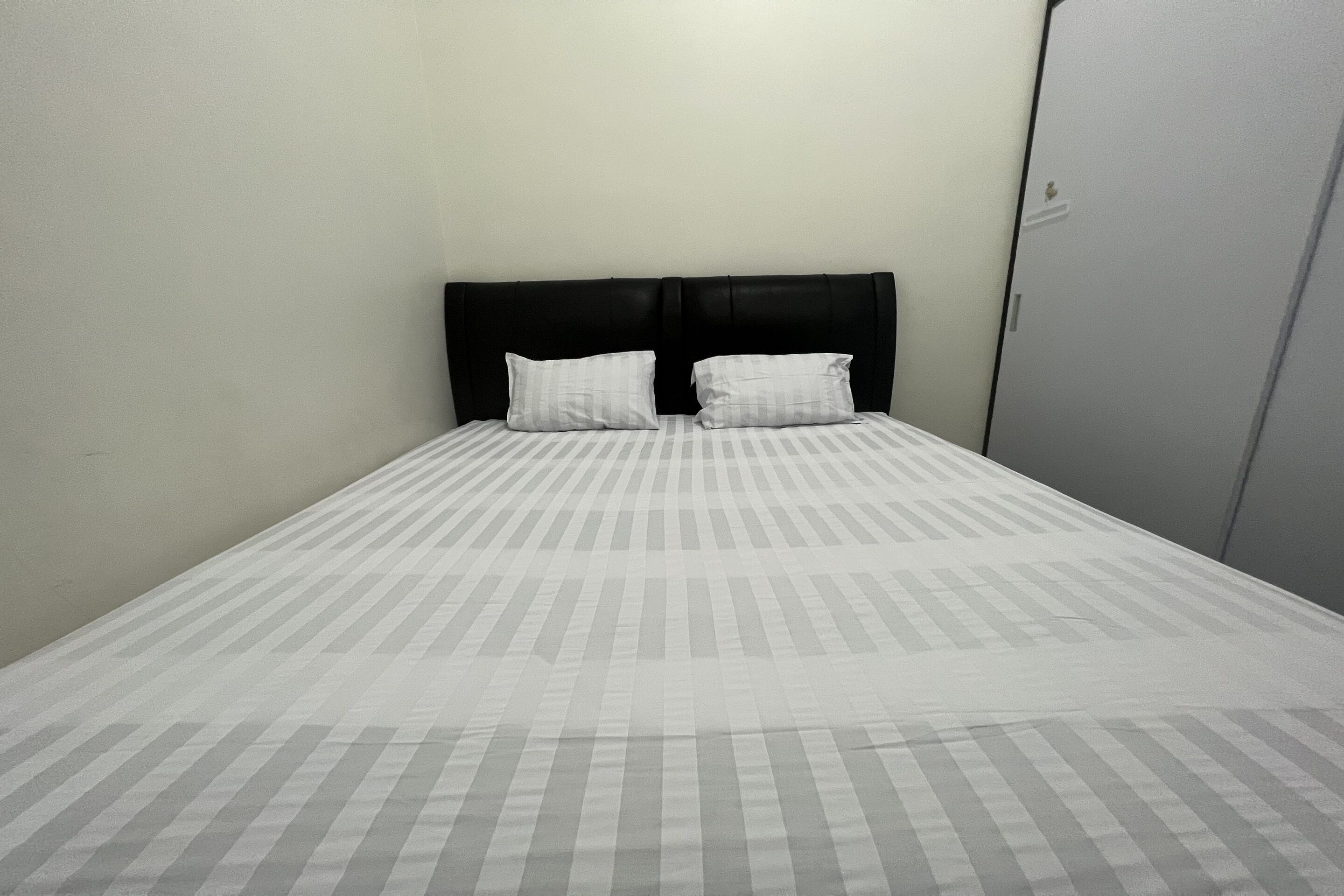 Un letto singolo con una testiera nera e due cuscini, una parete spoglia e una porta alla destra.