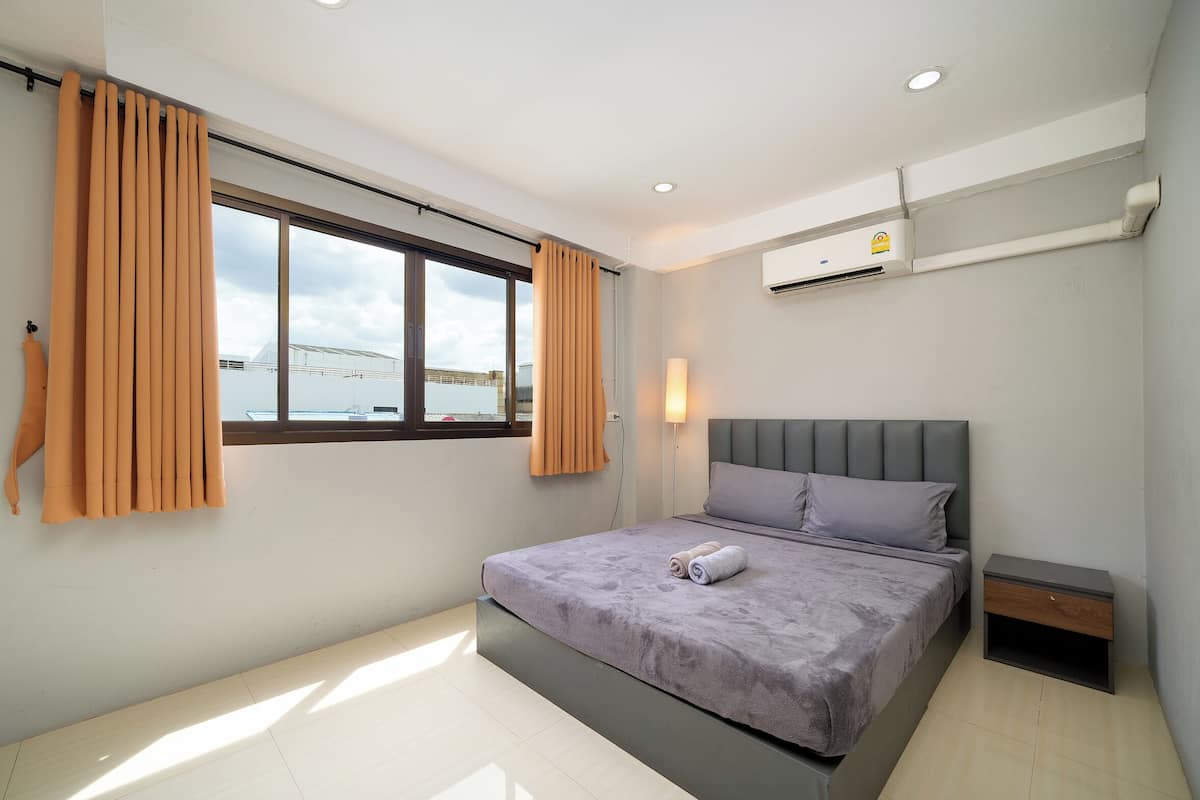 A modern hotel room with a large bed, a nightstand, a window with curtains, and an air conditioning unit.