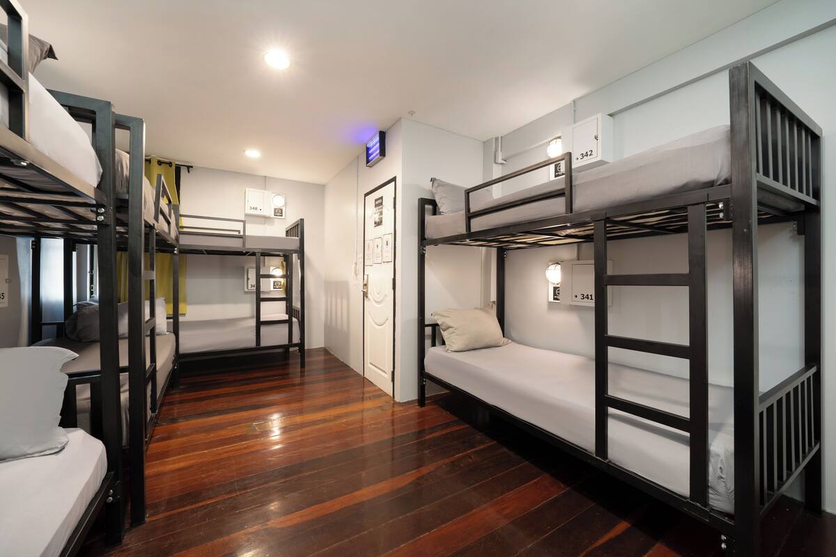 A dormitory-style room with bunk beds, wooden flooring, and a small desk with a chair.