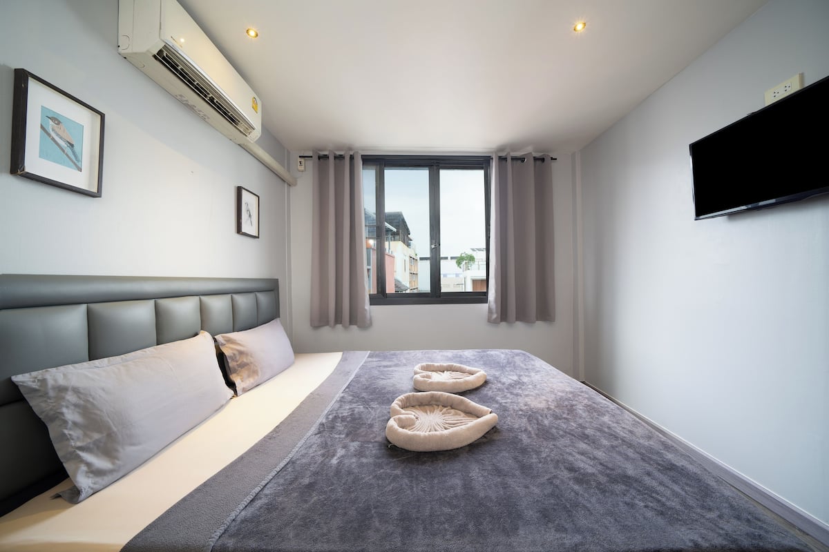 A modern hotel room with a large bed, a flat-screen TV, an air conditioning unit, and a window with curtains.
