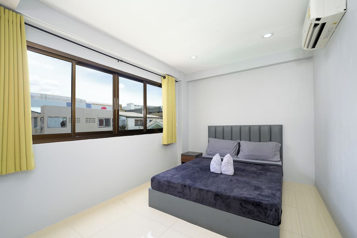 A modern bedroom with a large window, a bed with grey bedding, and an air conditioning unit on the wall.
