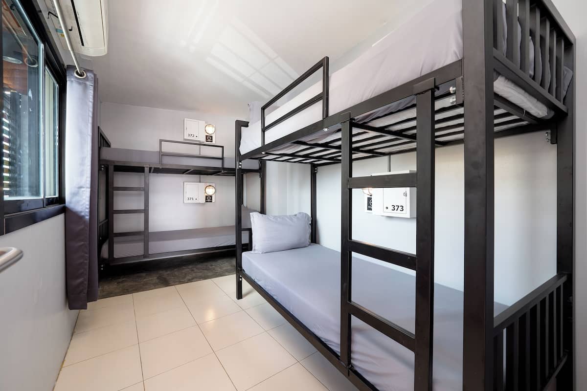 A dormitory room with bunk beds, a window with curtains, and a thermostat on the wall.