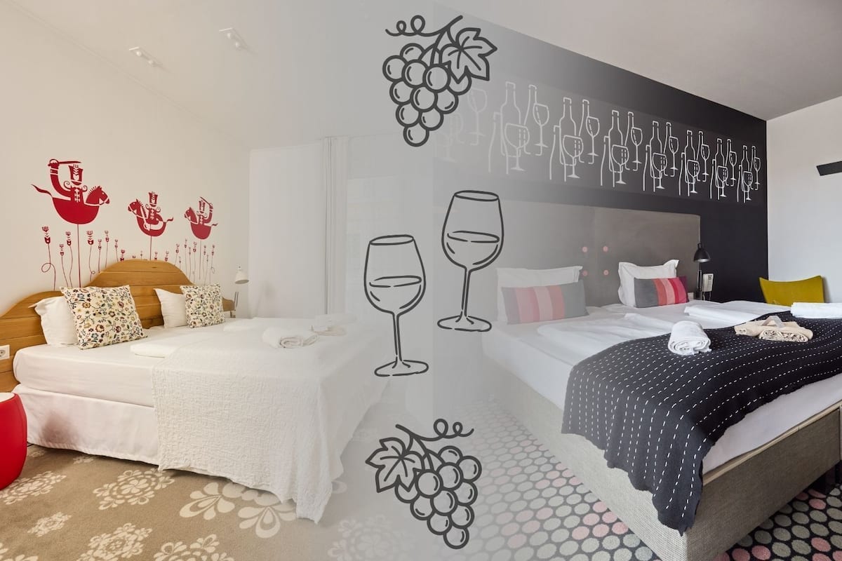 A hotel room with a bed, a red ottoman, a wall with grape and wine illustrations, and a patterned carpet.