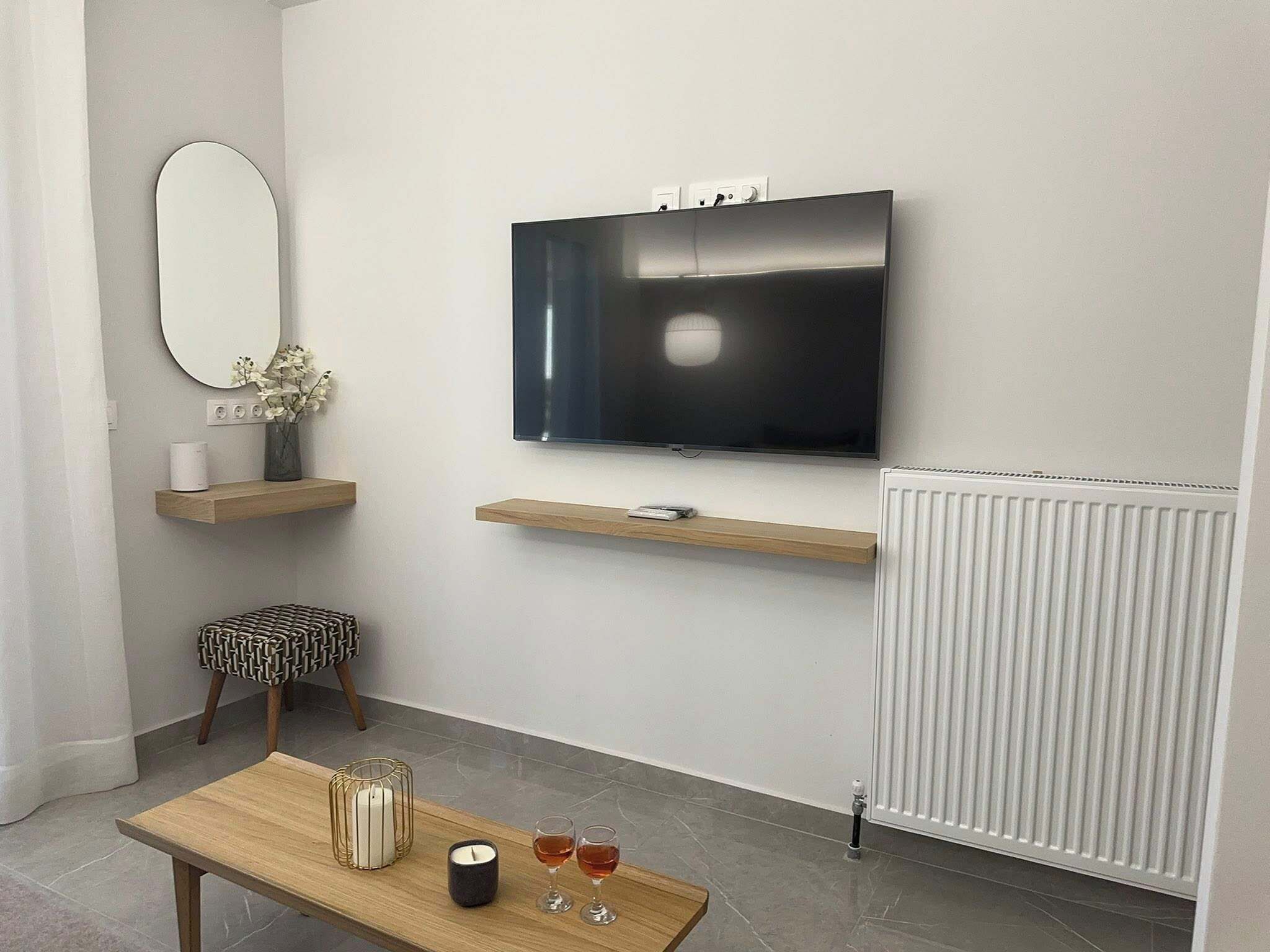 A modern living room with a flat-screen TV mounted on the wall, wooden shelves, a patterned ottoman, a small coffee table with candles and glasses, and a radiator.