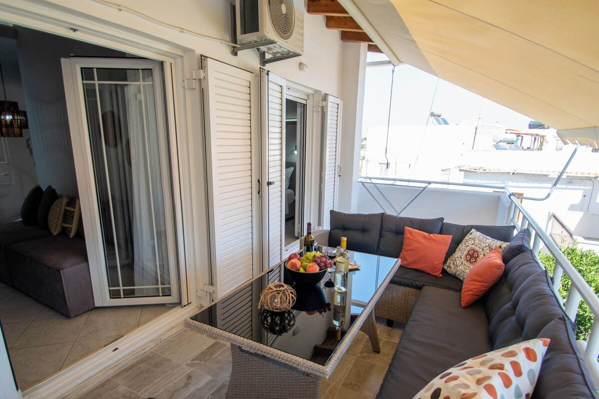 A balcony with a sofa, a glass table with a fruit bowl and bottles, and a sliding glass door leading to a room.