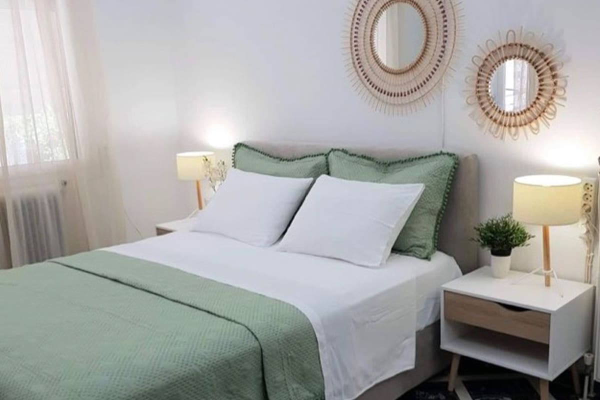 Uma cama bem arrumada com colcha verde e branca, duas almofadas brancas e duas luminárias de cabeceira sobre mesas de cabeceira brancas.