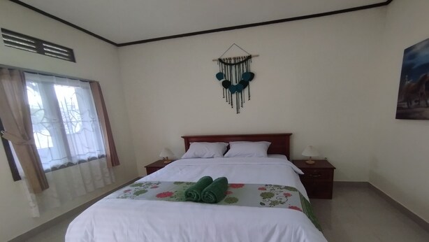 Una cama bien tendida con ropa blanca y toallas verdes, una mesita de noche con lámpara y un adorno en la pared.